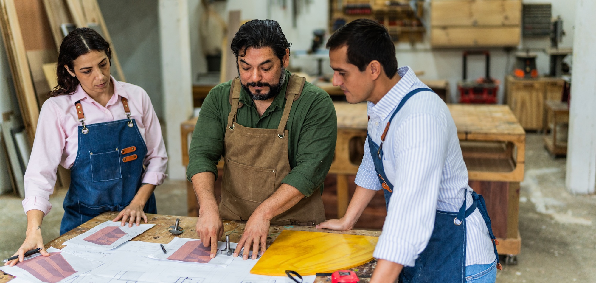 Three people are discussing plans in a wood-working shop